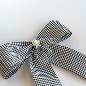 May include: A black and white gingham bow with a pearl accent. The bow is tied and ready to be used as a hair accessory or decorative element.
