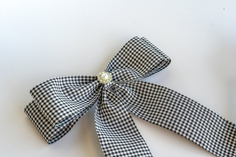 May include: A black and white gingham bow with a pearl accent. The bow is tied and ready to be used as a hair accessory or decorative element.