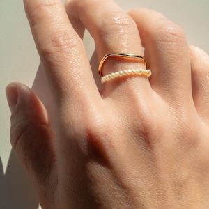 May include: Two gold rings on a finger. The top ring is a simple, curved band. The bottom ring is a delicate band of small pearls.