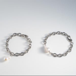 Puede incluir: Dos pulseras de cadena plateadas con eslabones grandes. Cada pulsera tiene un colgante de perla blanca de forma irregular. Las pulseras se muestran sobre una superficie blanca.