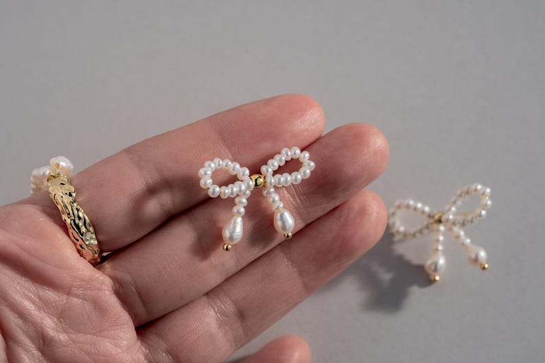 May include: A pair of pearl bow earrings with gold accents. The earrings are small and delicate, and they would make a great gift for a special occasion.