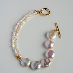 May include: A delicate bracelet featuring two strands of small, round pearls and a row of larger, iridescent coin pearls. The bracelet has gold-colored accents and a toggle clasp. The pearls have a soft, lustrous sheen.