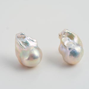 Puede incluir: Dos perlas barrocas iridiscentes, con base blanco crema y destellos rosas, verdes y azules. Las perlas tienen formas irregulares y una superficie lisa y brillante. Ideales para la creación de joyas.