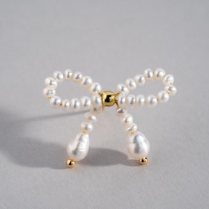 May include: A white pearl bow tie brooch with a gold center. The bow is made of small pearls and the center is a gold bead.