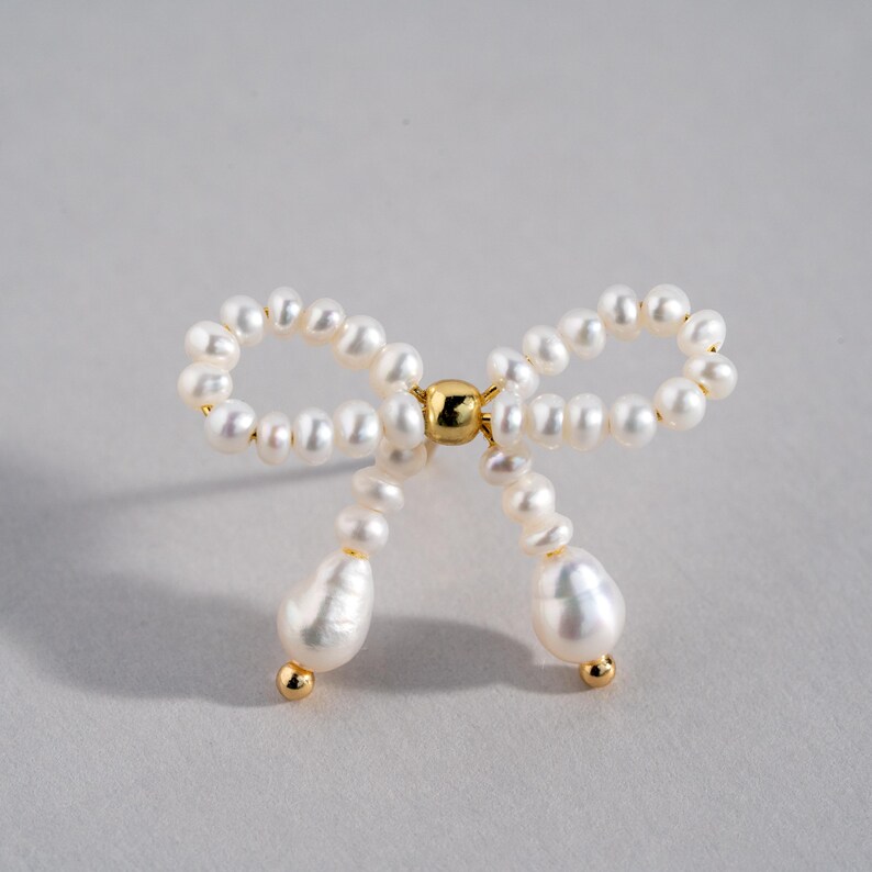 May include: A white pearl bow tie brooch with a gold center. The bow is made of small pearls and the center is a gold bead.