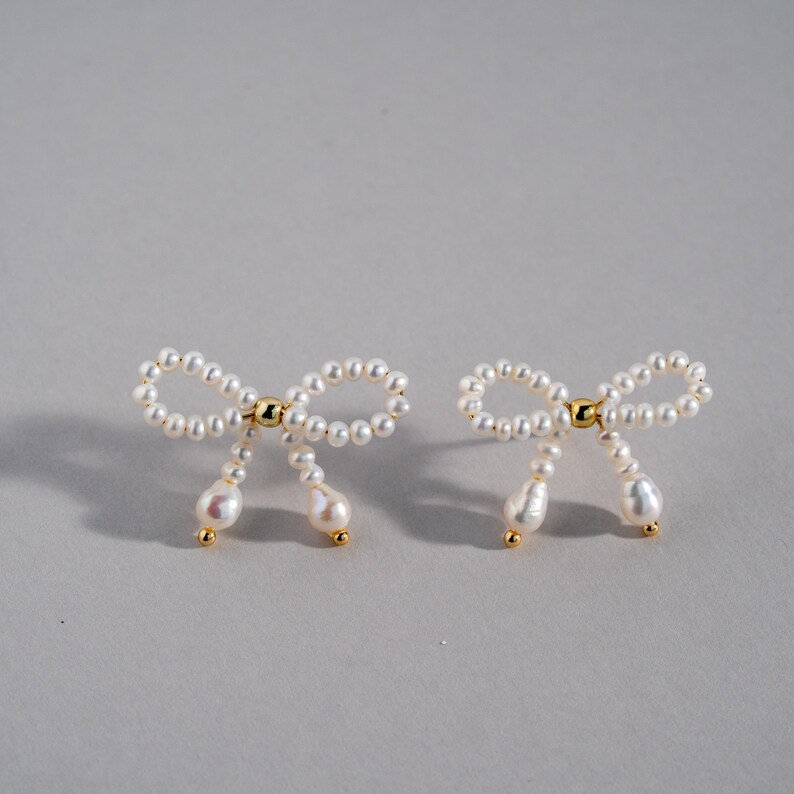 May include: A pair of pearl earrings shaped like bows. The earrings are made of small white pearls and have a gold accent in the center of each bow.