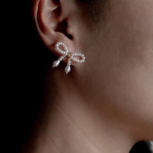 May include: A pair of pearl and gold bow earrings. The earrings are made of small white pearls and have a gold bow in the center. The earrings are shown on a person's ear.