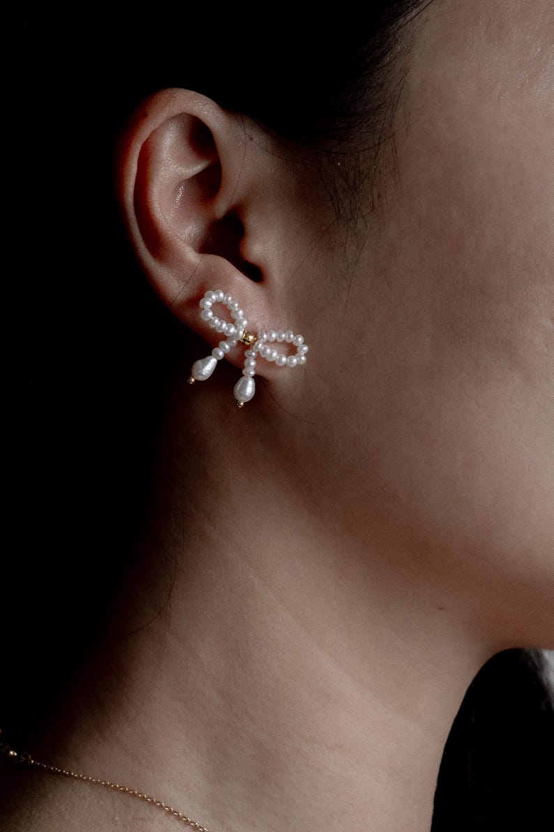 May include: A pair of pearl and gold bow earrings. The earrings are made of small white pearls and have a gold bow in the center. The earrings are shown on a person's ear.