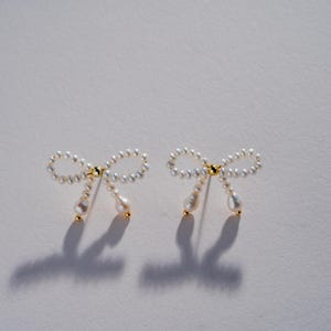 May include: A pair of pearl bow earrings with a gold accent. The earrings are made of small white pearls and have a delicate bow shape.