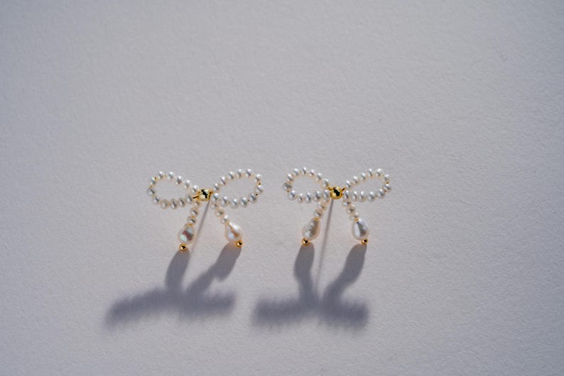 May include: A pair of pearl bow earrings with a gold accent. The earrings are made of small white pearls and have a delicate bow shape.
