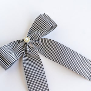 May include: A black and white gingham bow with a pearl and gold accent. The bow is tied with a long tail.
