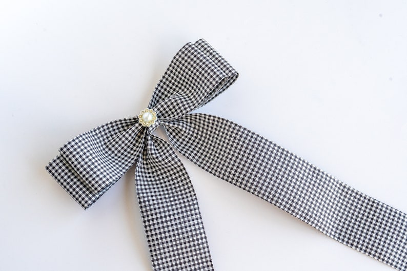 May include: A black and white gingham bow with a pearl and gold accent. The bow is tied with a long tail.