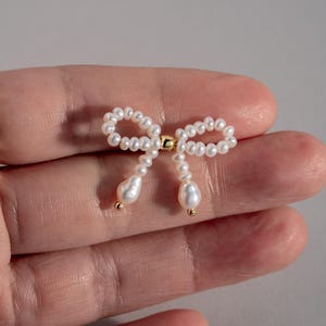 May include: A delicate pearl bow earring with a gold accent. The bow is made of small white pearls and the accent is a small gold bead.