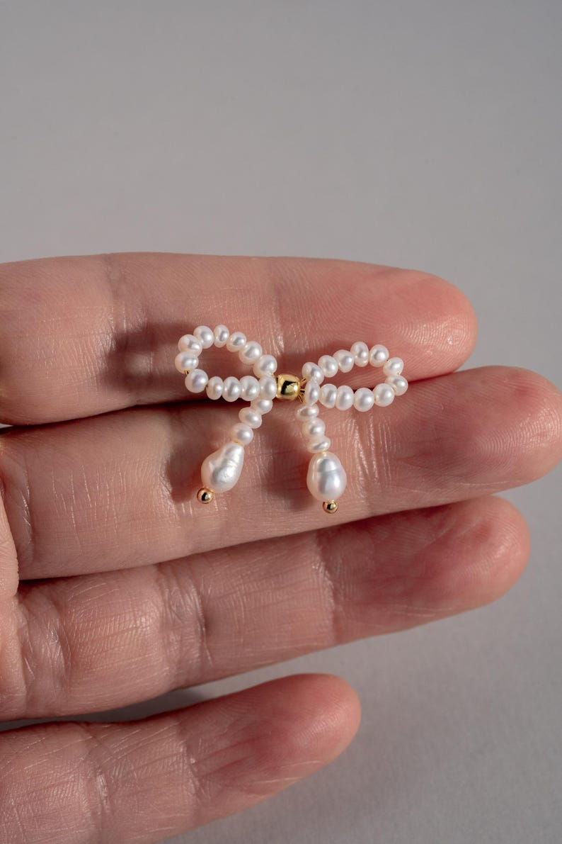 May include: A delicate pearl bow earring with a gold accent. The bow is made of small white pearls and the accent is a small gold bead.