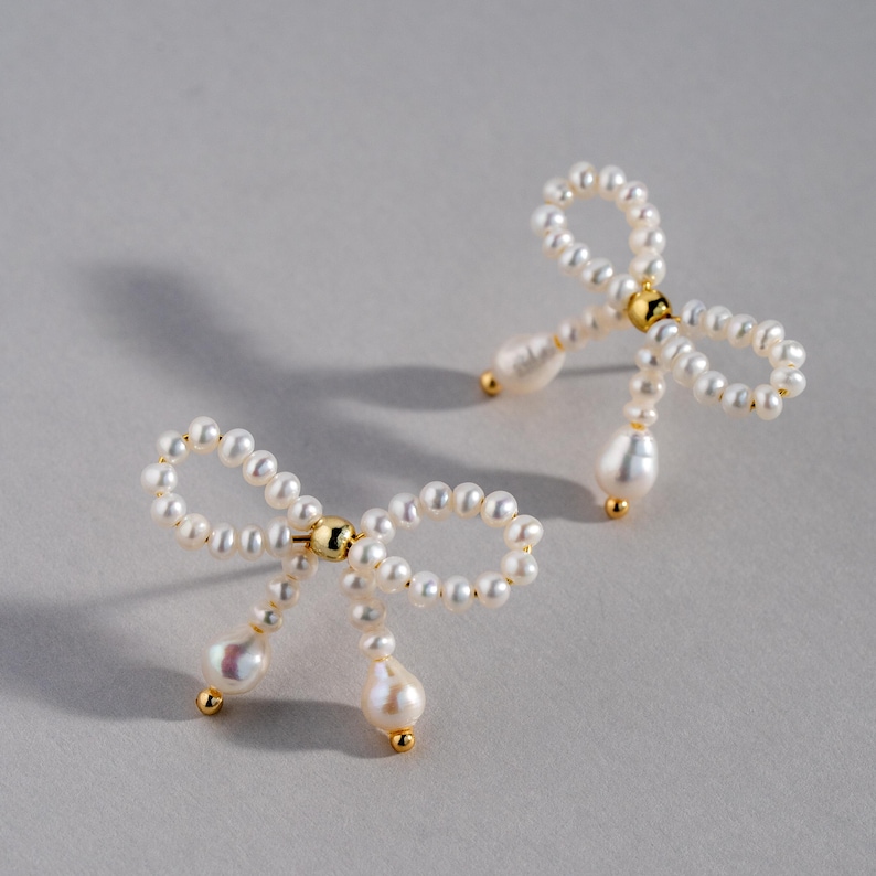 May include: A pair of pearl earrings shaped like bows with dangling pearl drops. The earrings are made of white pearls and gold metal.