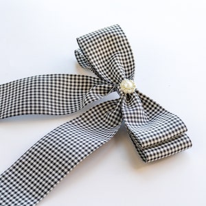 May include: A black and white gingham ribbon bow with a pearl accent. The bow is tied in a large, loose knot and has long tails.