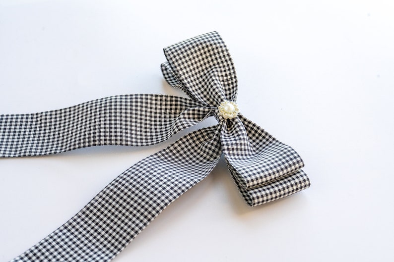 May include: A black and white gingham ribbon bow with a pearl accent. The bow is tied in a large, loose knot and has long tails.
