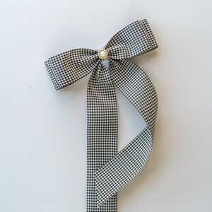 May include: A black and white gingham ribbon bow with a pearl accent. The bow is tied with a long tail.