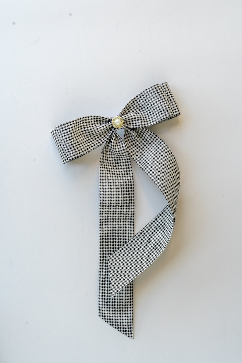 May include: A black and white gingham ribbon bow with a pearl accent. The bow is tied with a long tail.
