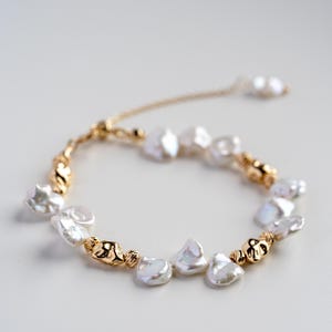 Puede incluir: Una delicada pulsera con perlas blancas irregulares y cuentas doradas. La pulsera está ensartada en una fina cadena dorada, con un cierre deslizante ajustable. El diseño es simple pero elegante, adecuado para diversas ocasiones.