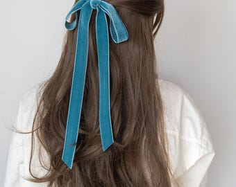 May include: A teal velvet hair bow tied in a bow, with long, flowing tails. The bow is attached to long, wavy brown hair. The person is wearing a white shirt. The background is a plain, light gray.
