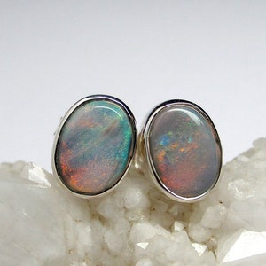 May include: A pair of silver stud earrings with oval-shaped opal gemstones. The opals have a rainbow of colors, including green, blue, and red.