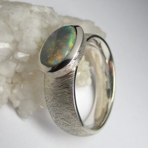 May include: A silver ring with a textured band and an oval-shaped opal gemstone. The opal has a green, blue, and black iridescent sheen.