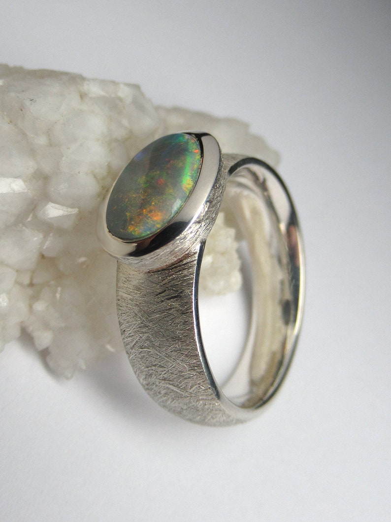 May include: A silver ring with a textured band and an oval-shaped opal gemstone. The opal has a green, blue, and black iridescent sheen.