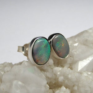 May include: A pair of silver stud earrings with oval-shaped opal gemstones. The opals have a white base with green and brown iridescent flashes.