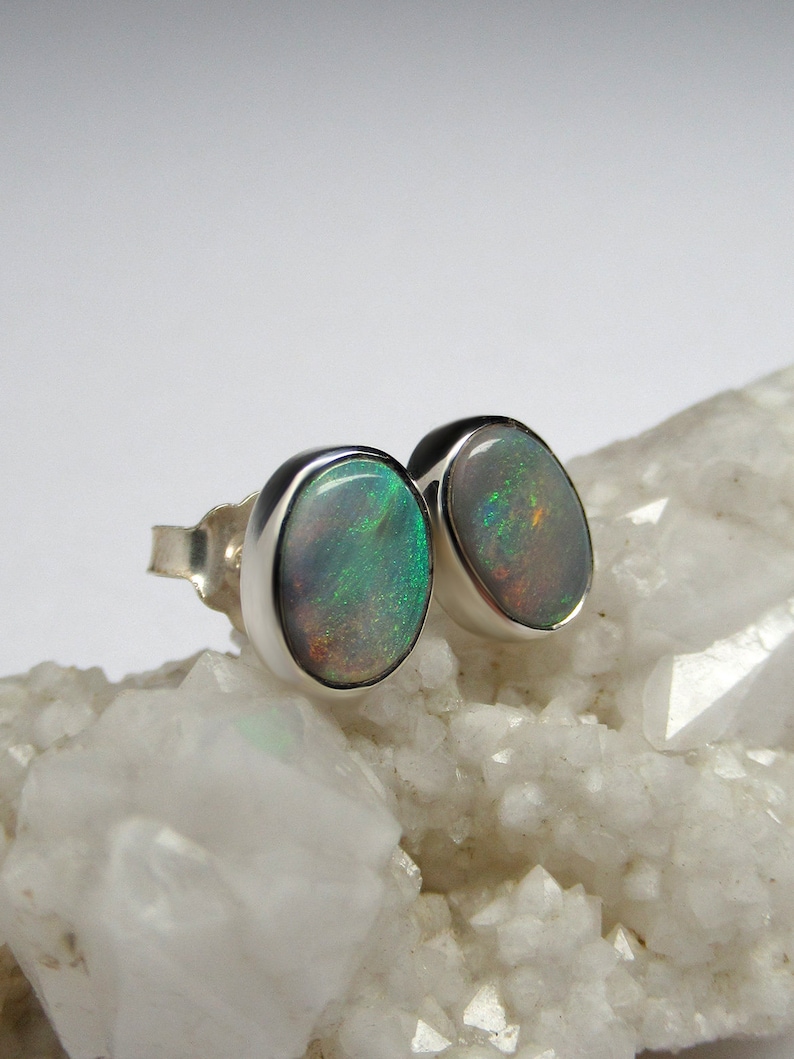 May include: A pair of silver stud earrings with oval-shaped opal gemstones. The opals have a white base with green and brown iridescent flashes.