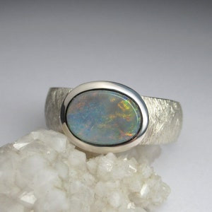 May include: A silver ring with a textured band and a round opal gemstone set in a bezel setting. The opal has a blue, green, and orange iridescent sheen.