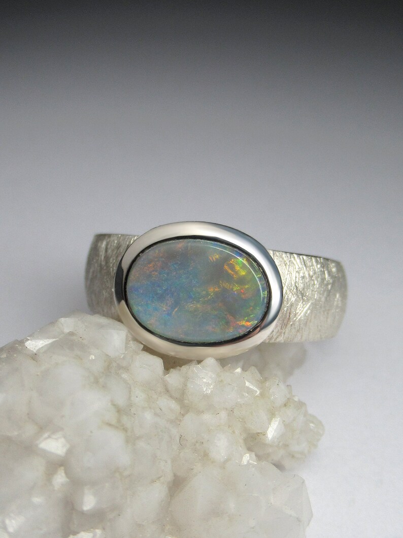 May include: A silver ring with a textured band and a round opal gemstone set in a bezel setting. The opal has a blue, green, and orange iridescent sheen.
