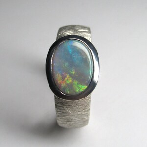 May include: A silver ring with a textured band and an oval-shaped opal gemstone. The opal has a blue, green, and yellow iridescent sheen.