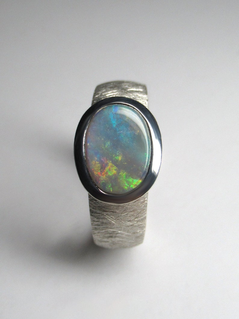 May include: A silver ring with a textured band and an oval-shaped opal gemstone. The opal has a blue, green, and yellow iridescent sheen.