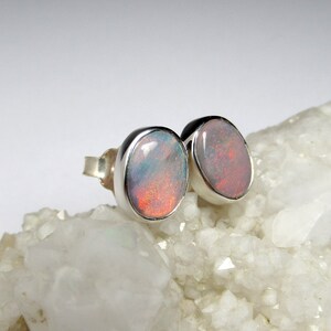 May include: A pair of silver stud earrings with oval-shaped opal gemstones. The opals have a white, pink, and orange iridescent sheen.