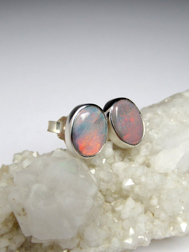 May include: A pair of silver stud earrings with oval-shaped opal gemstones. The opals have a white, pink, and orange iridescent sheen.