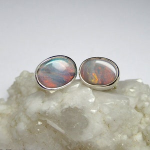 May include: A pair of silver earrings with oval-shaped opal gemstones. The opals have a rainbow of colors, including pink, blue, green, and orange.