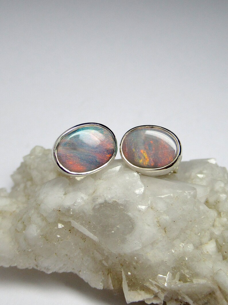 May include: A pair of silver earrings with oval-shaped opal gemstones. The opals have a rainbow of colors, including pink, blue, green, and orange.