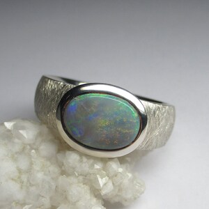 May include: A silver ring with a textured band and a large oval opal gemstone. The opal has a green, blue, and black iridescent sheen.
