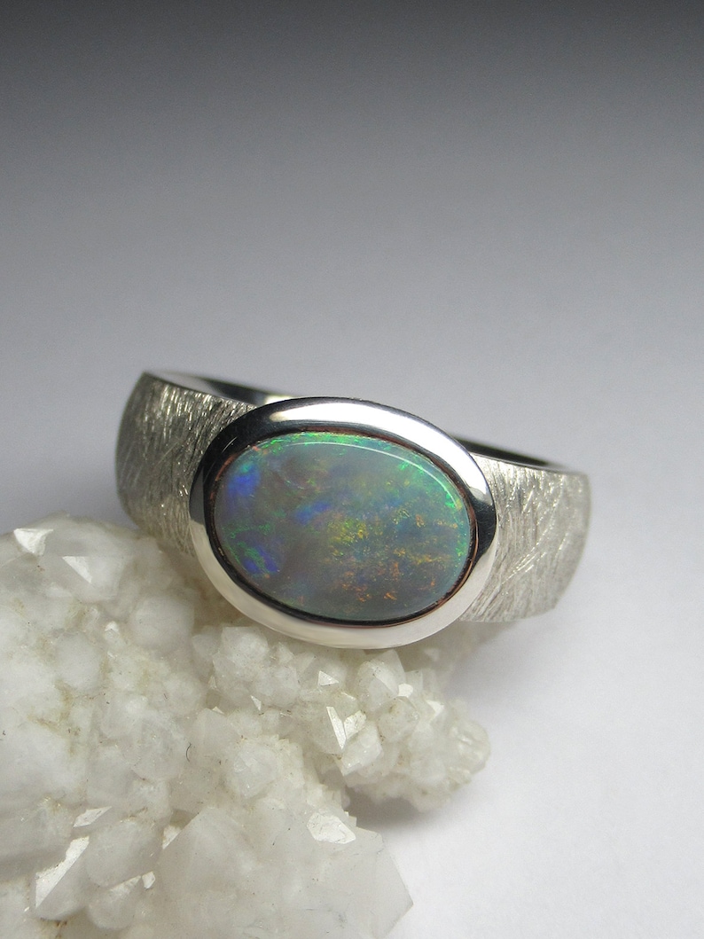 May include: A silver ring with a textured band and a large oval opal gemstone. The opal has a green, blue, and black iridescent sheen.