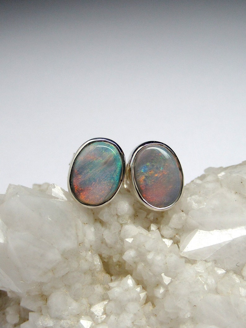 May include: A pair of silver stud earrings with oval-shaped opal gemstones. The opals have a rainbow of colors, including green, blue, and red.