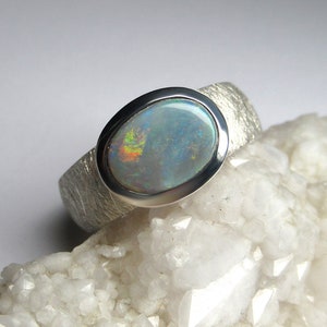 May include: A silver ring with a textured band and a large oval opal gemstone. The opal has a white base with flashes of blue, green, and red.