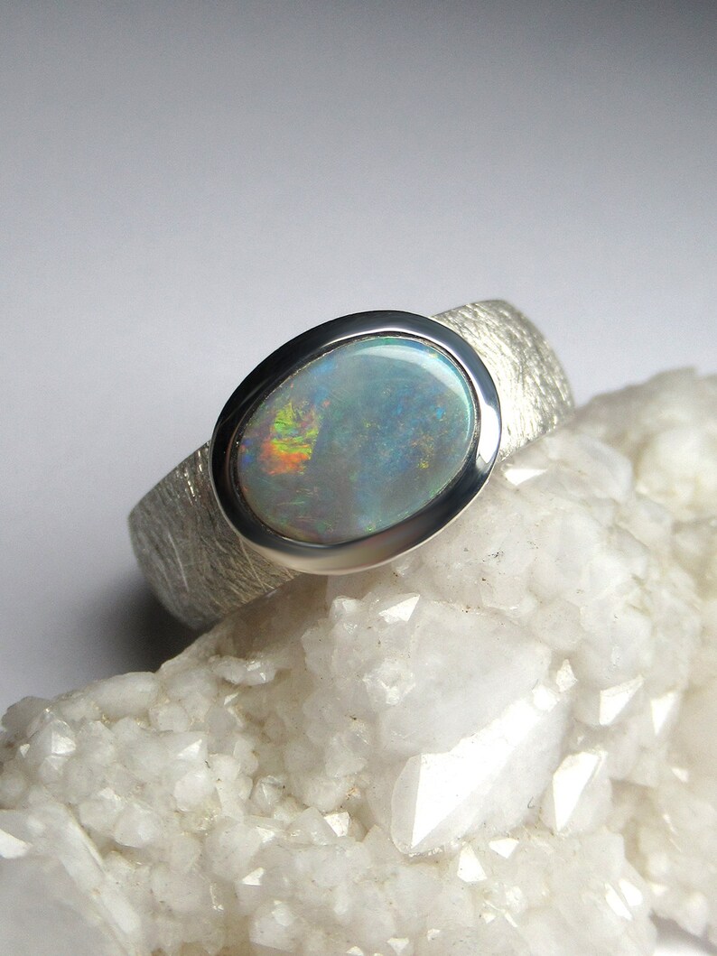 May include: A silver ring with a textured band and a large oval opal gemstone. The opal has a white base with flashes of blue, green, and red.