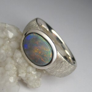 May include: A silver ring with a textured band and a large oval opal gemstone. The opal has a dark grey base with flashes of blue, green, and orange.