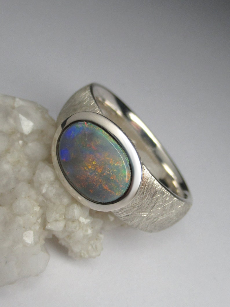 May include: A silver ring with a textured band and a large oval opal gemstone. The opal has a dark grey base with flashes of blue, green, and orange.
