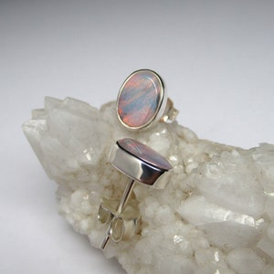 May include: A pair of silver stud earrings with oval-shaped opal gemstones. The opals have a pink, blue, and white iridescent sheen.