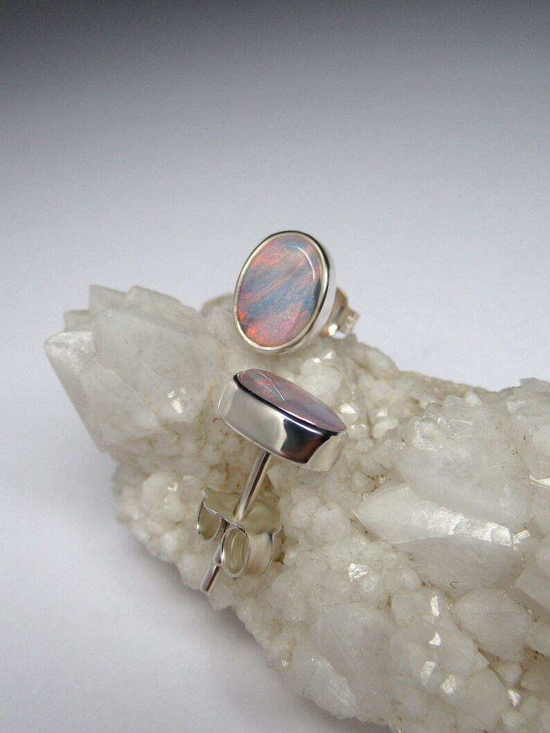 May include: A pair of silver stud earrings with oval-shaped opal gemstones. The opals have a pink, blue, and white iridescent sheen.