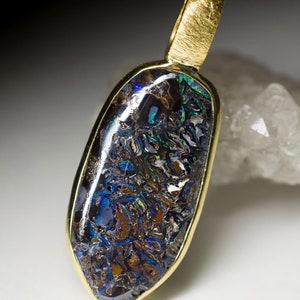 May include: A gold pendant featuring a dark blue and brown opal with iridescent flashes of green and blue. The opal is set in a gold bezel and has a smooth, oval shape.