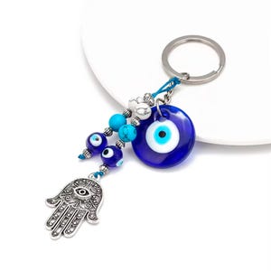 May include: A keychain featuring a blue evil eye charm, a silver Hamsa hand pendant, and decorative beads in shades of blue and white. The keychain has a silver keyring and is designed to ward off negative energy.