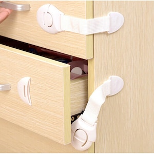 Può includere: Serrature di sicurezza per bambini di colore bianco per armadi e cassetti. Le serrature hanno un cinturino bianco e un chiavistello in plastica bianca. Le serrature sono installate su un armadio in legno.